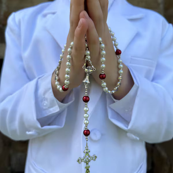 Person wearing a white shirt holding a rosary with red and gold beads.